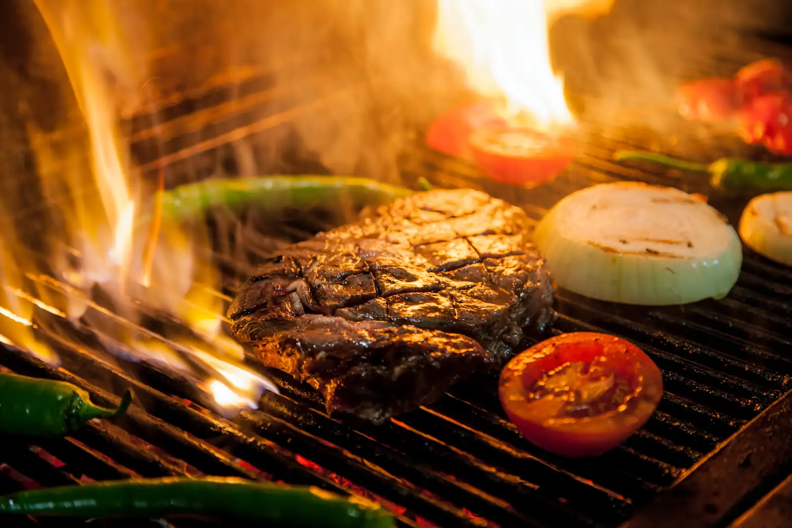 Steak searing over an open flame alongside grilled onion, tomato and peppers